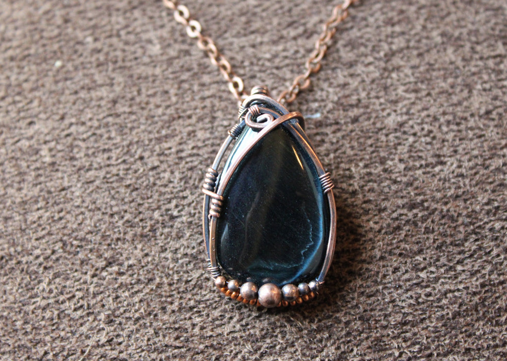 Tear drop shaped dark blue tigers eye stone wrapped in copper set on a brown textured background. 