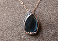 Tear drop shaped dark blue tigers eye stone wrapped in copper set on a brown textured background. 
