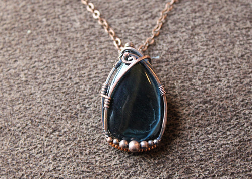Tear drop shaped dark blue tigers eye stone wrapped in copper set on a brown textured background. 