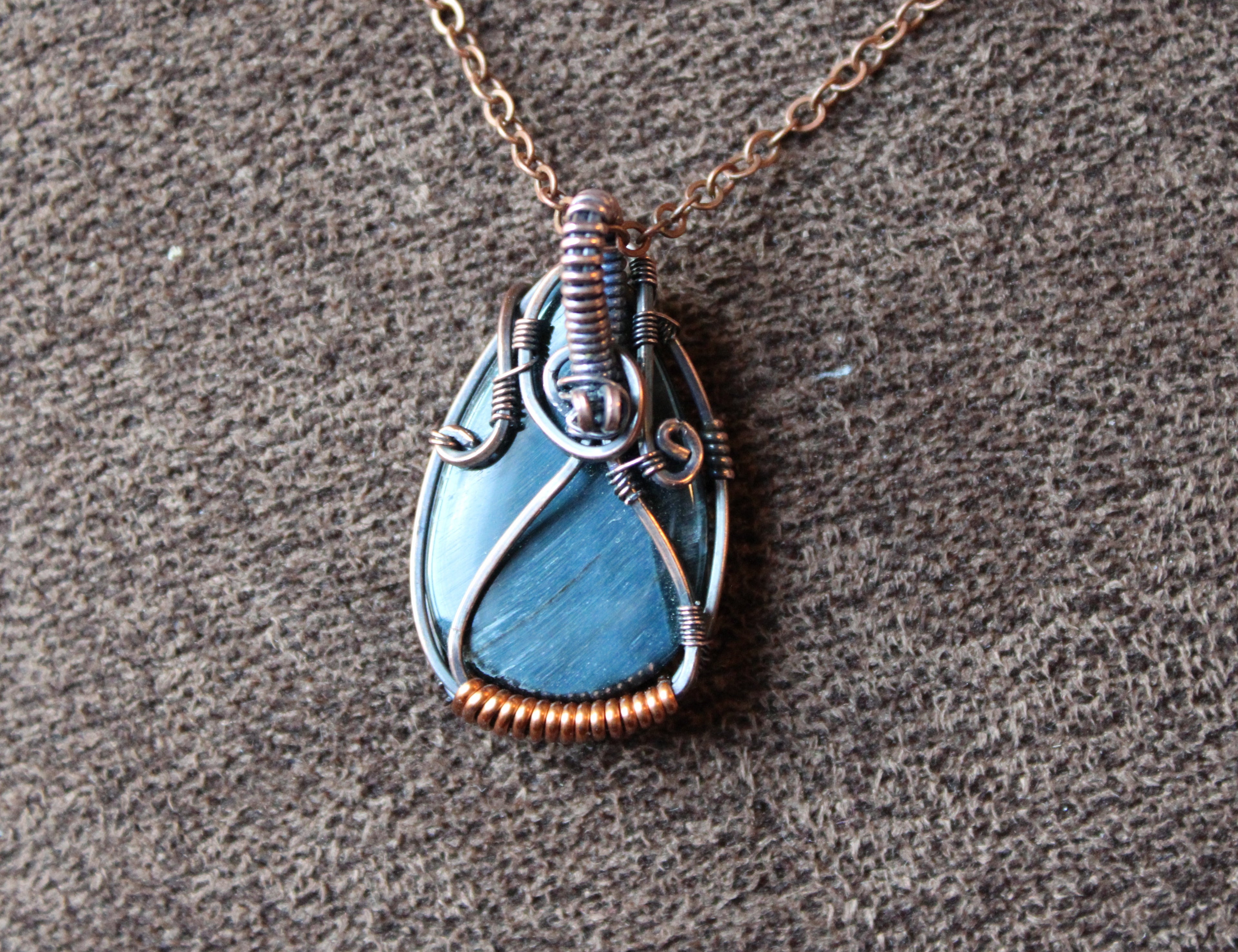 Tear drop shaped dark blue tigers eye stone wrapped in copper set on a brown textured background. 