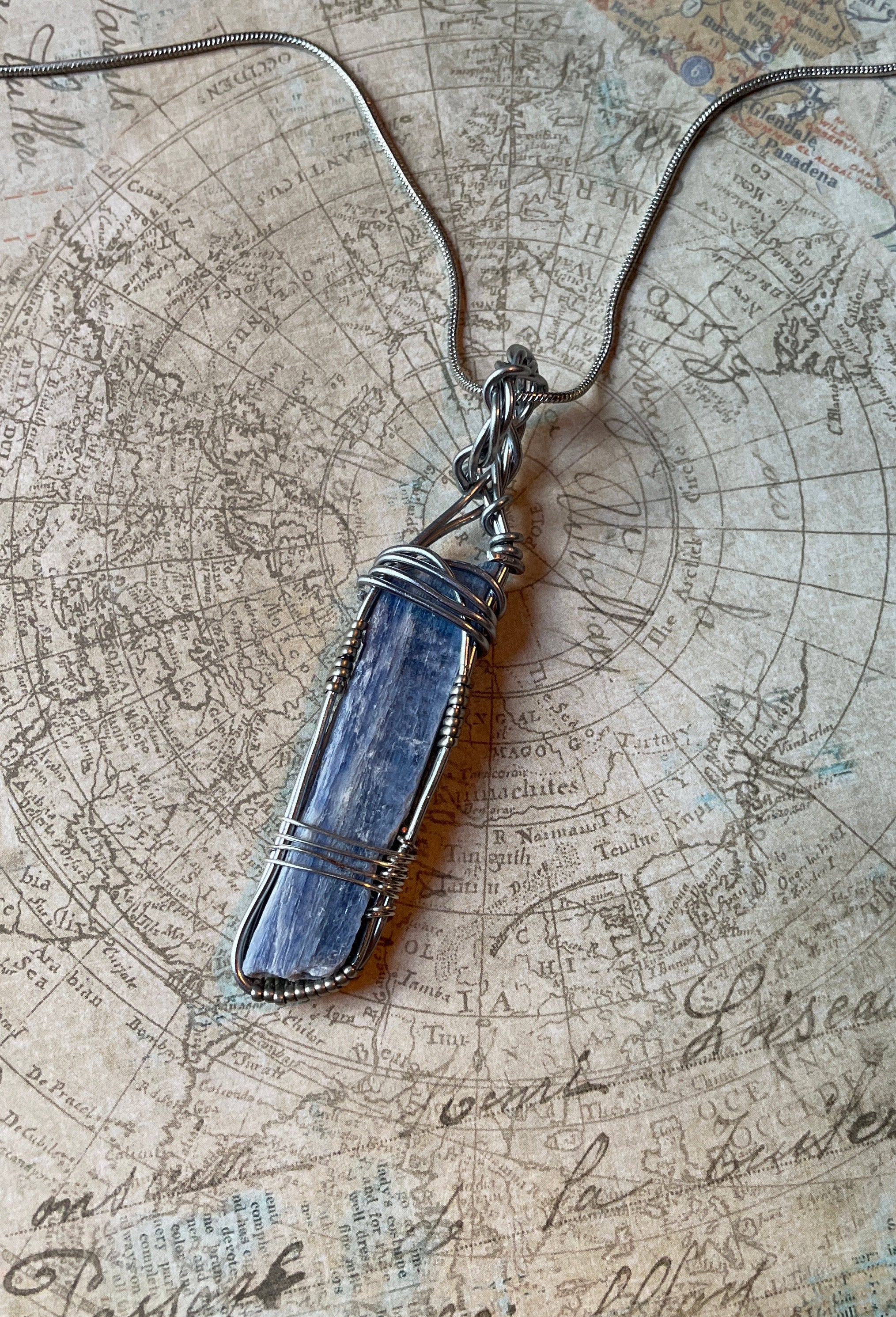 A silver toned wire-wrapped pendant necklace featuring a blue-gray crystal, laid flat on an antique-style map background.