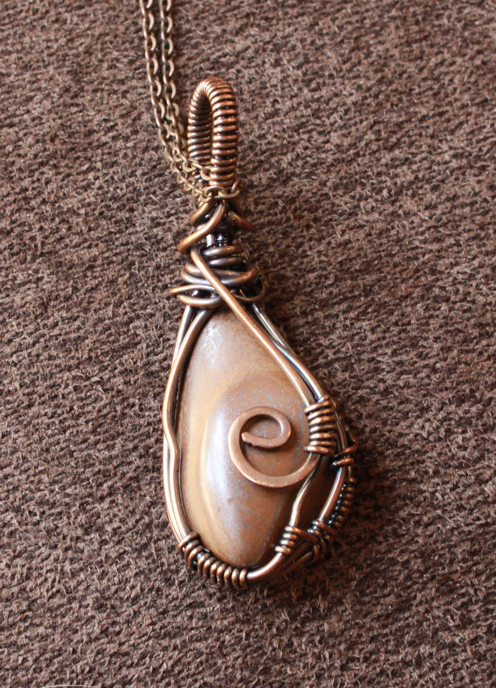 A teardrop-shaped wire-wrapped pendant necklace featuring a natural cowrie shell with a spiral shell detail, set in copper-toned wire, shown on a textured brown background.