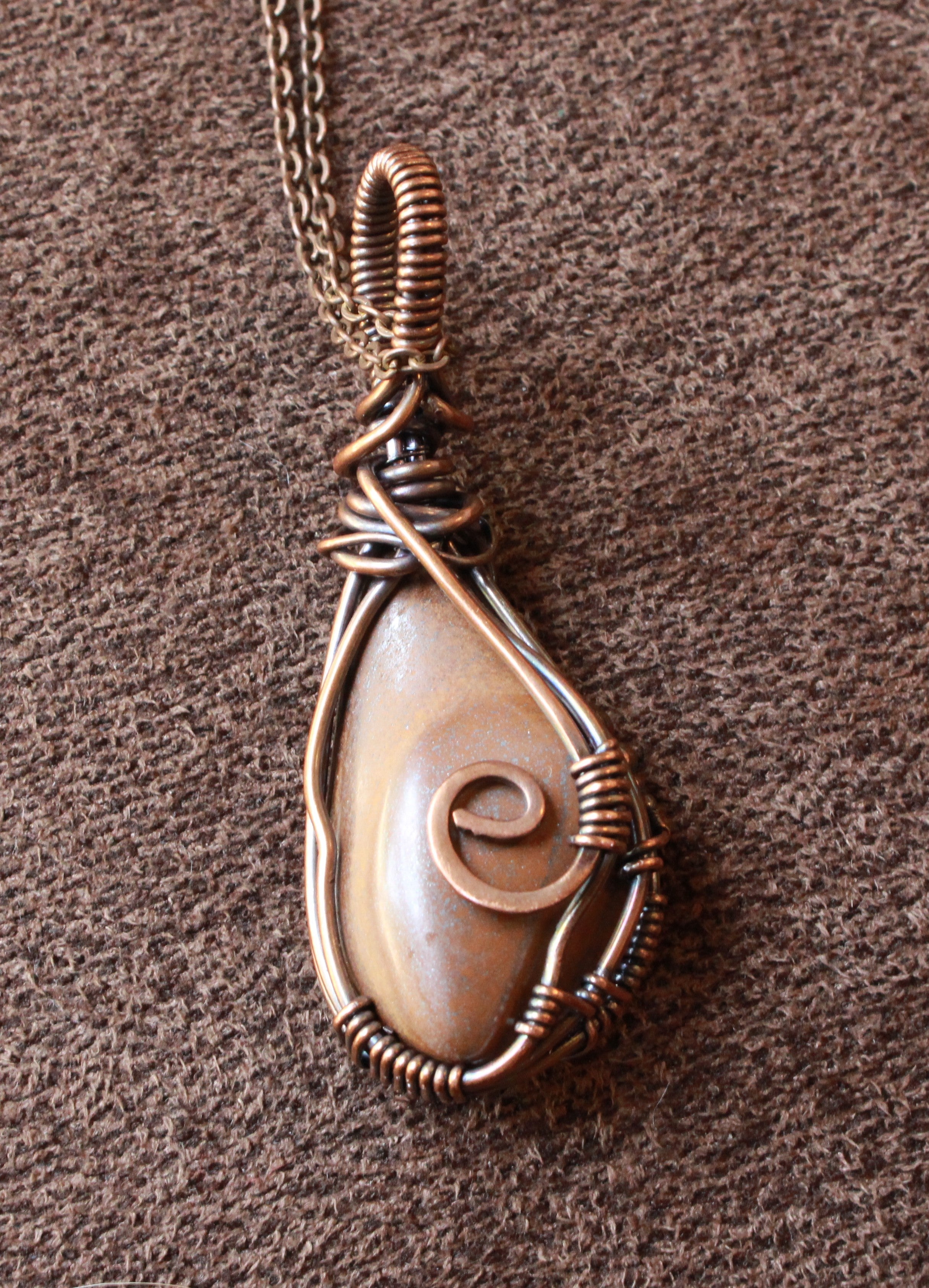A teardrop-shaped wire-wrapped pendant necklace featuring a natural cowrie shell with a spiral shell detail, set in copper-toned wire, shown on a textured brown background.