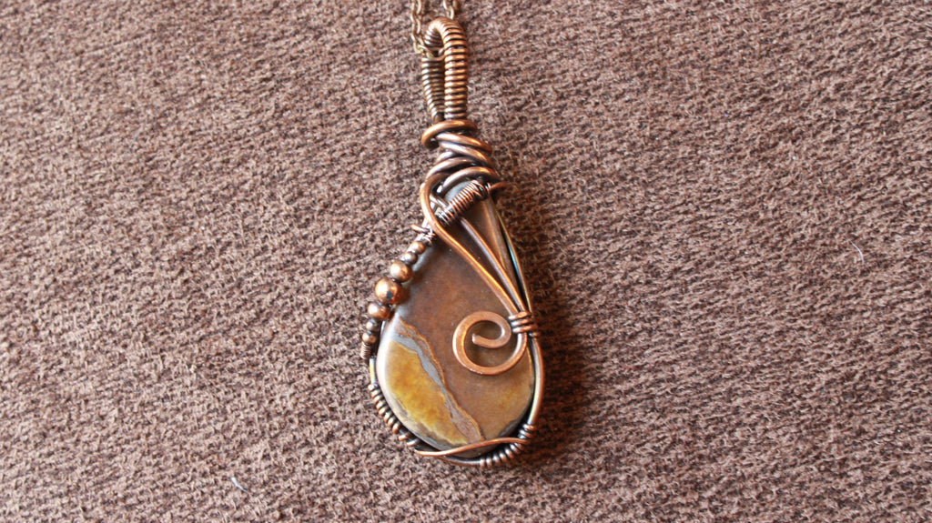 A handmade teardrop pendant featuring a natural brown and gold stone wrapped in copper-toned wire, resting on a textured brown fabric background.