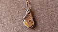 A handmade teardrop pendant featuring a natural brown and gold stone wrapped in copper-toned wire, resting on a textured brown fabric background.