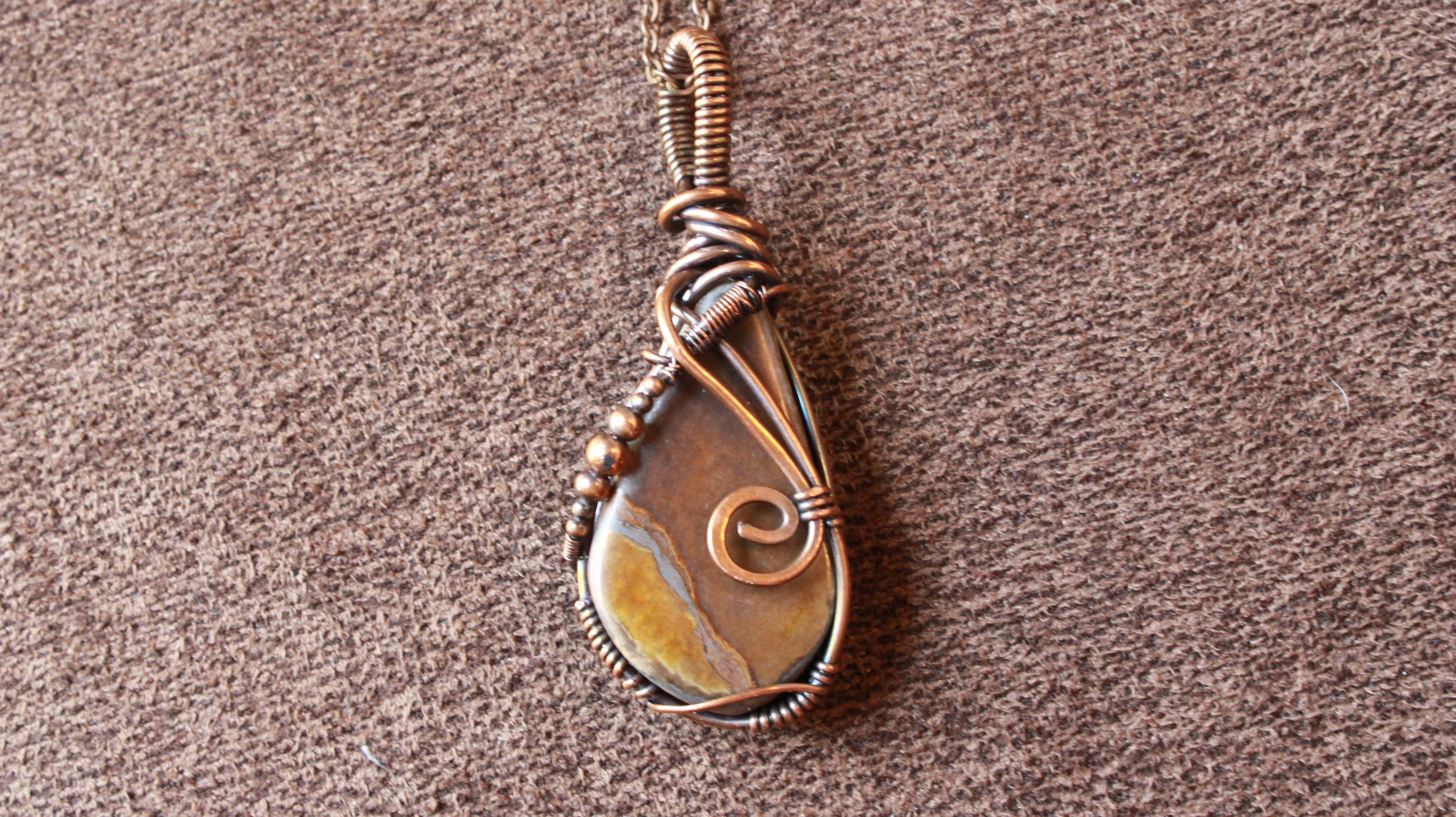 A handmade teardrop pendant featuring a natural brown and gold stone wrapped in copper-toned wire, resting on a textured brown fabric background.